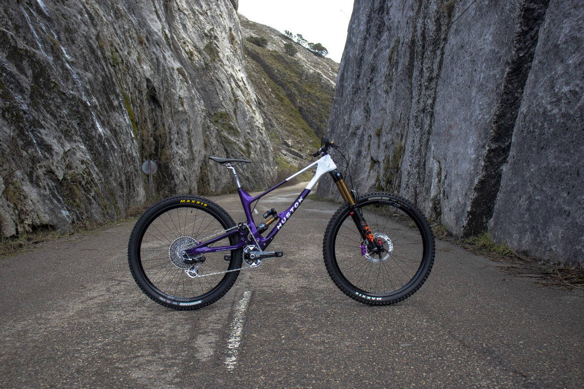 A purple Hustok mountain bike positioned in a narrow rocky canyon, with steep stone walls on either side and a paved path running through the center.