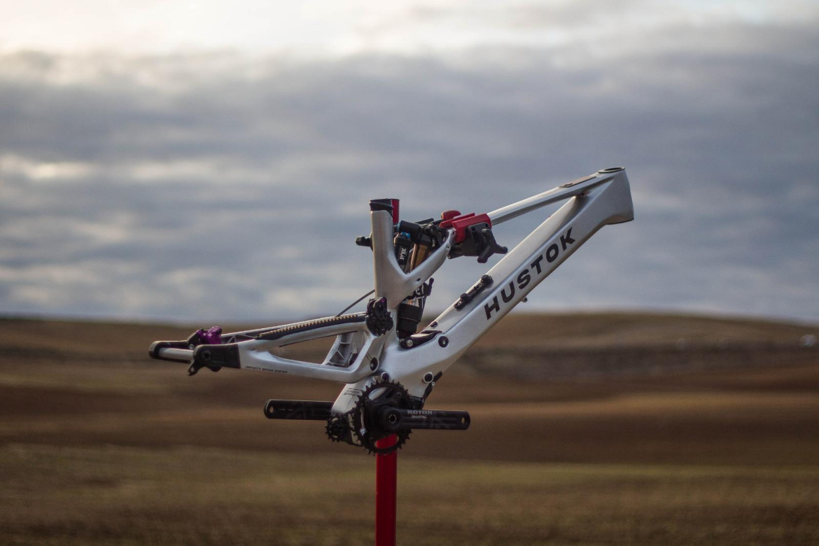 A mountain bike frame from the brand Hustok is mounted on a repair stand, set against a cloudy sky and an open field. The frame features a white finish with black and red components.
