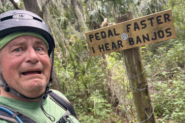 A person on a biking trail with a concerned expression, wearing a helmet and a green shirt, stands next to a wooden sign that reads "PEDAL FASTER, I HEAR BANJOS." The background features lush greenery and hanging moss, suggesting a forested area. Loyce E. Harpe Park mountain bike trail.