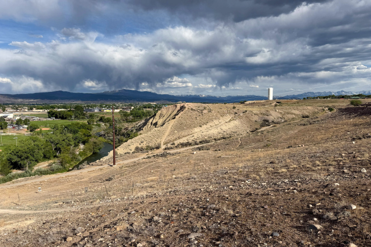 A panoramic view of a dry, hilly landscape with a winding river at the base. In the distance, a small town is visible, along with green trees and open fields. Dark storm clouds gather in the sky above the mountains, creating a dramatic backdrop. A water tower can be seen on the horizon, indicating human presence in the area.