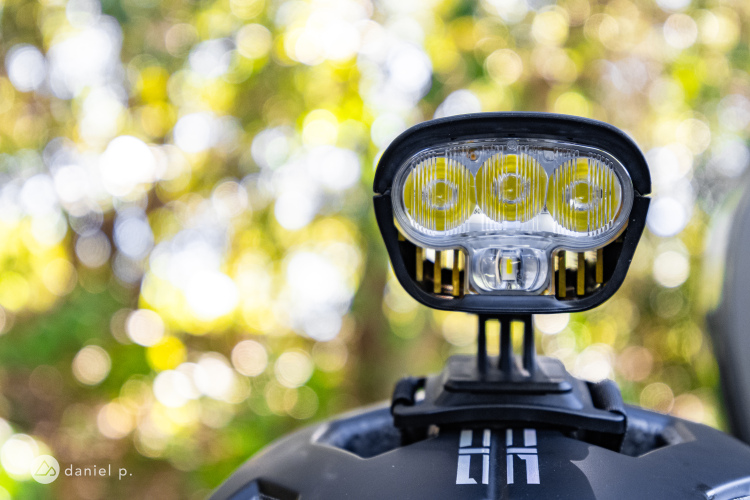 A close-up view of a bike light mounted on a helmet, with a blurred background of greenery. The light features three bright LED bulbs encased in a transparent cover, mounted securely on the helmet. The overall image conveys a sense of outdoor adventure and safety.