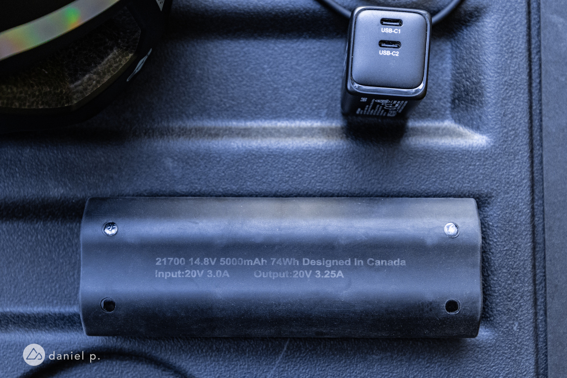 Close-up image showing a black battery labeled "21700 14.8V 5000mAh 74Wh Designed In Canada" alongside a charging unit with two USB ports (USB-C1 and USB-C2) and specifications for input and output. The background features a textured surface.