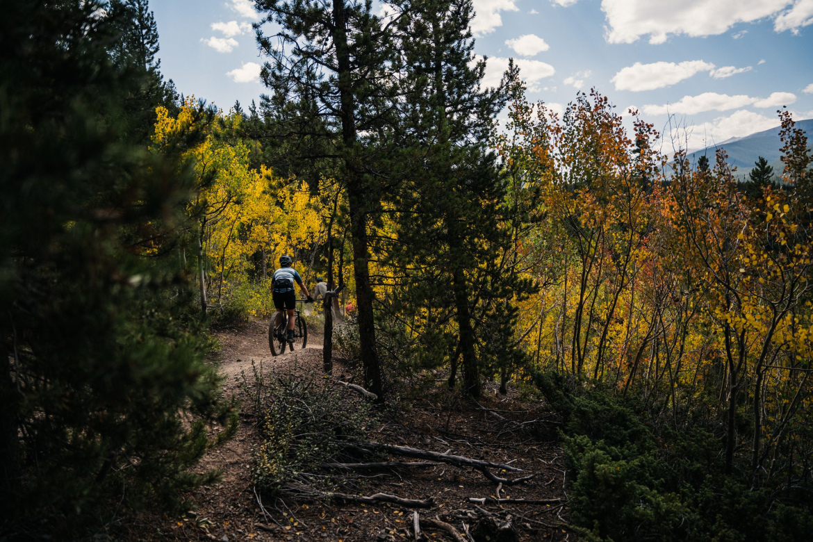 A cyclist riding Aspen Alley in Breckenridge, Colorado, surrounded by autumn foliage, with vibrant yellow and orange leaves on the trees. The scene is set in a forest with a mix of coniferous and deciduous trees under a partly cloudy sky.