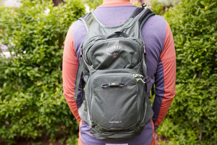 A person standing outdoors with their back to the camera, wearing a colorful long-sleeve shirt and a green Osprey Raptor 14 backpack. The background features lush green foliage.