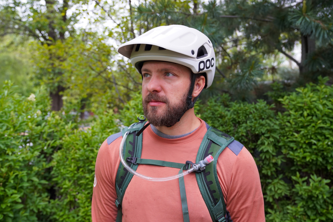 A man wearing a white POC Cularis Pure cycling helmet and a green backpack with a hydration tube stands in front of lush green foliage. He has a beard and is dressed in an orange long-sleeve shirt.