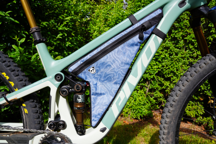 Close-up of a mountain bike frame featuring a blue patterned frame bag, shock absorbers, and a rubber tire. The background includes green foliage, highlighting the bike's design and detail.