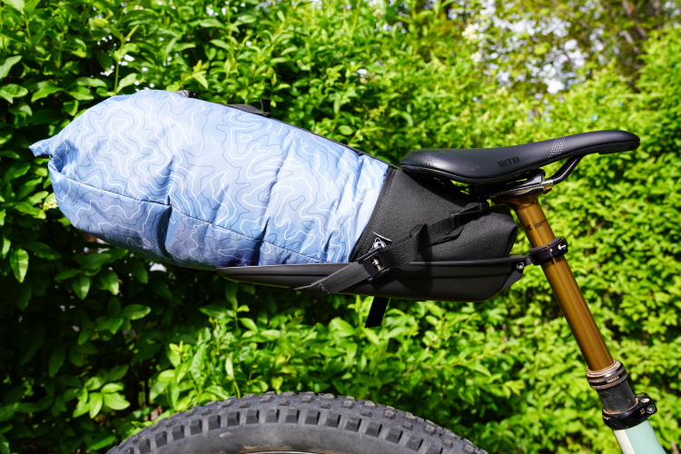 A blue patterned bicycle seat pack attached to a bike's saddle, with a backdrop of green foliage and a visible bike tire in the foreground.