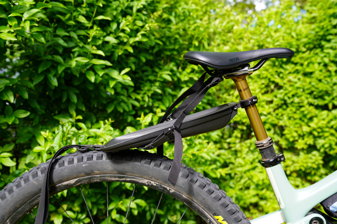 Close-up of a mountain bike seat with a black rear fender attached, set against a background of lush green foliage. The bike features a gold seat post and a thick, knobby tire, showcasing a combination of functionality and outdoor aesthetics.
