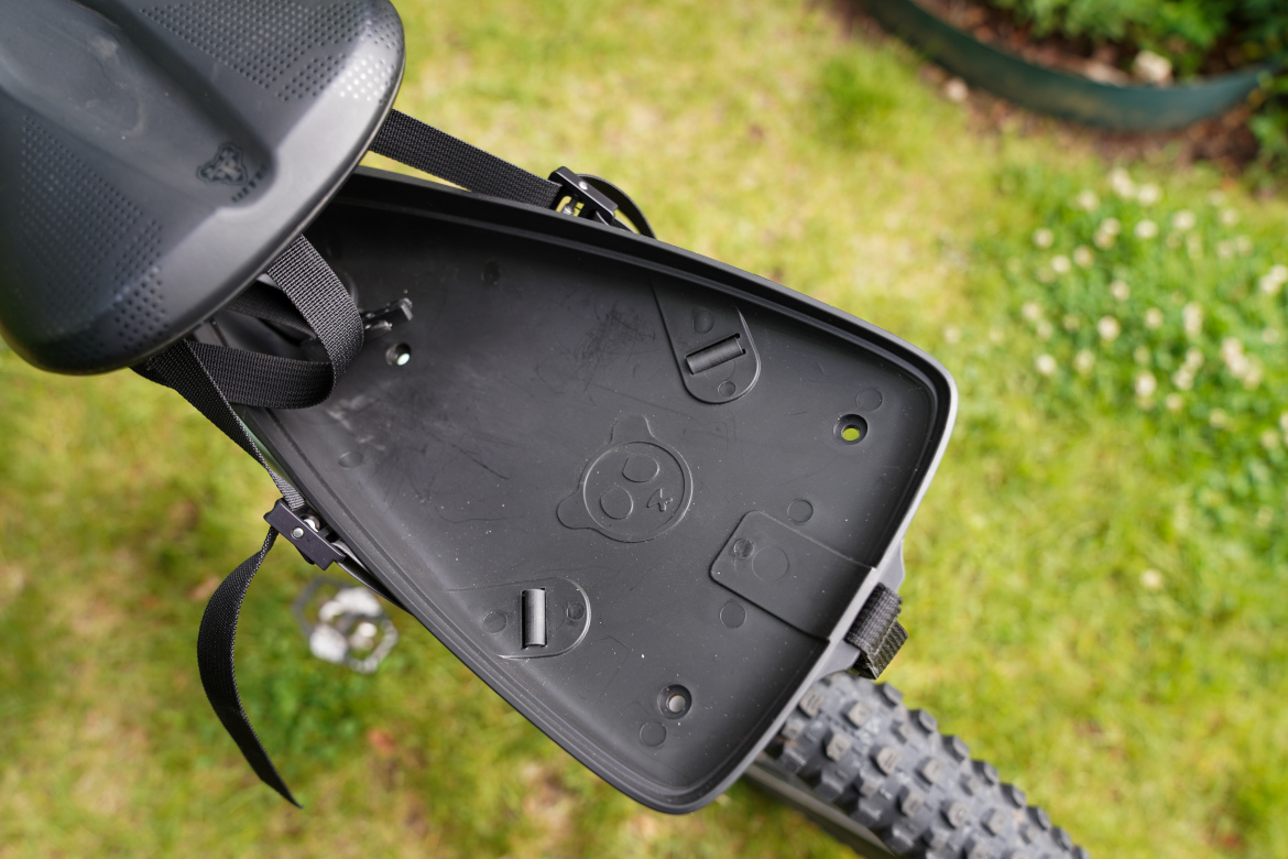 Close-up view of the underside of a bike seat, showing a textured black surface and fastening straps, with a grassy background. The seat features a small design detail at the center and is attached to a bicycle with visible pedal and tire tread in the frame.