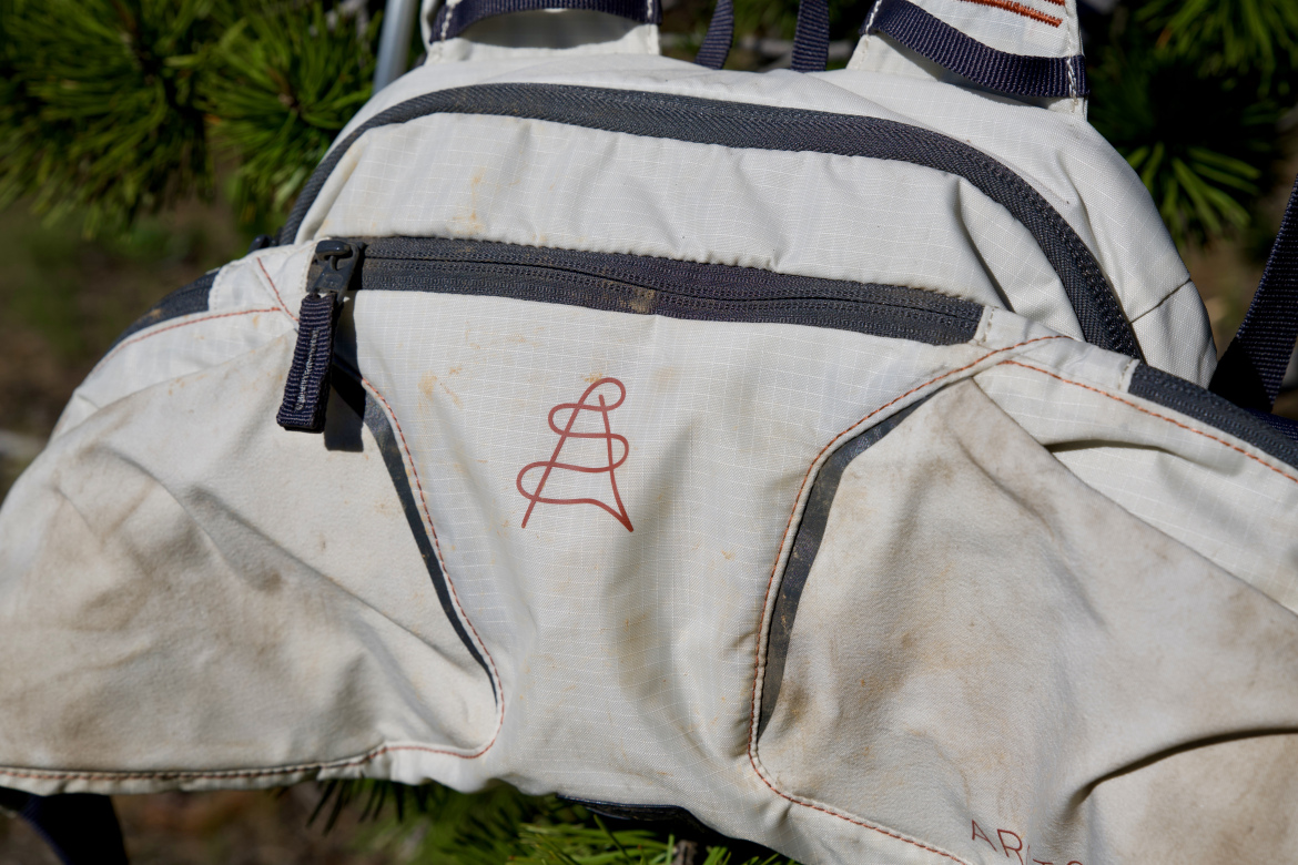 A close-up of a light-colored backpack with a logo and visible dirt marks, resting against a tree branch. The backpack features dark accents, zippers, and a geometric design on the front.