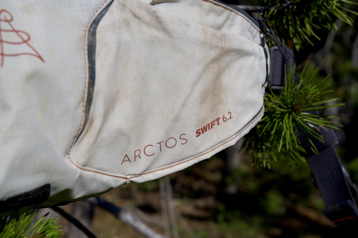 Close-up of a white backpack featuring the text "ARCTOS SWIFT 6.2," with some dirt marks visible. The backpack is hanging on a tree branch, surrounded by greenery.