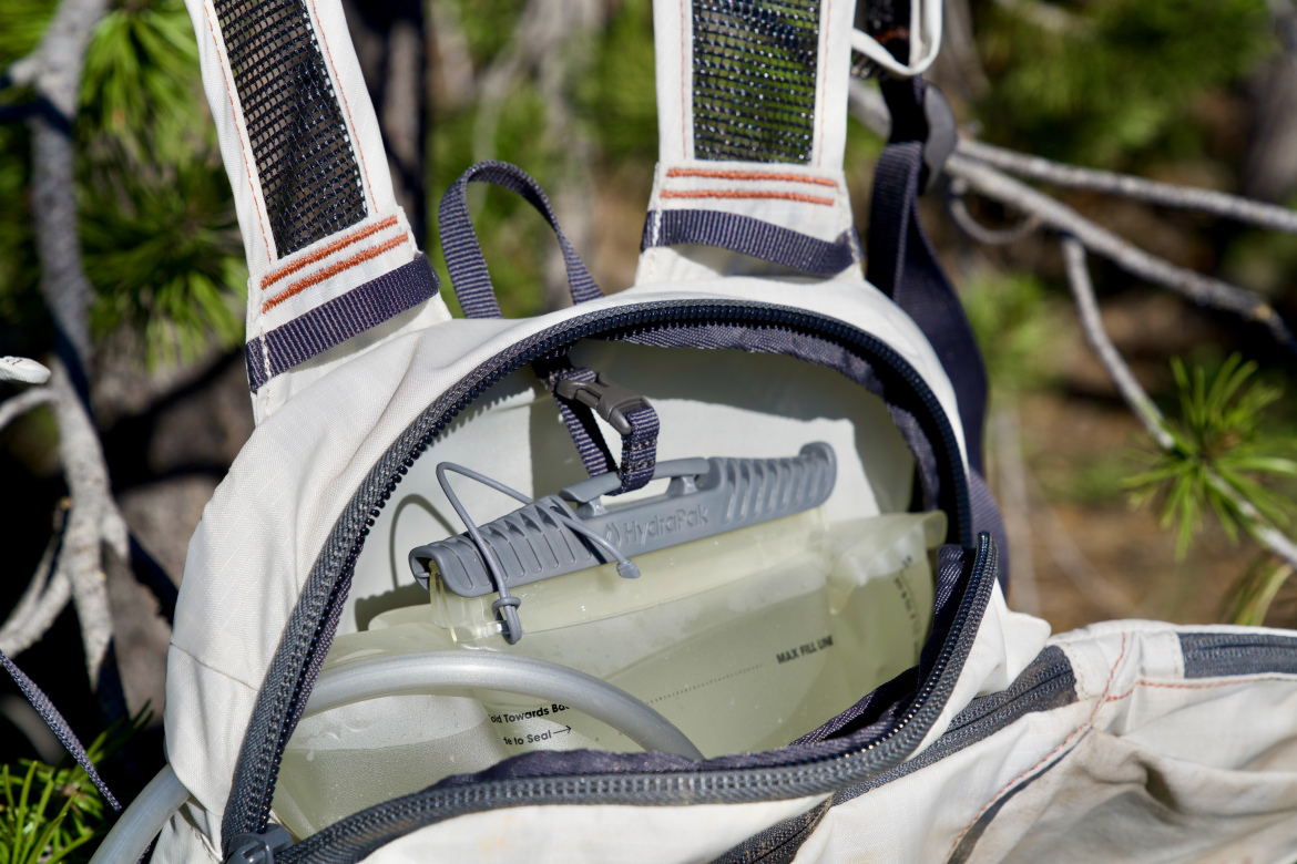 A close-up view of an open hydration pack featuring a clear water reservoir and a tube ready for use. The pack's exterior is light-colored with dark accents and features adjustable straps, with the background showing blurred pine tree branches, suggesting an outdoor setting.