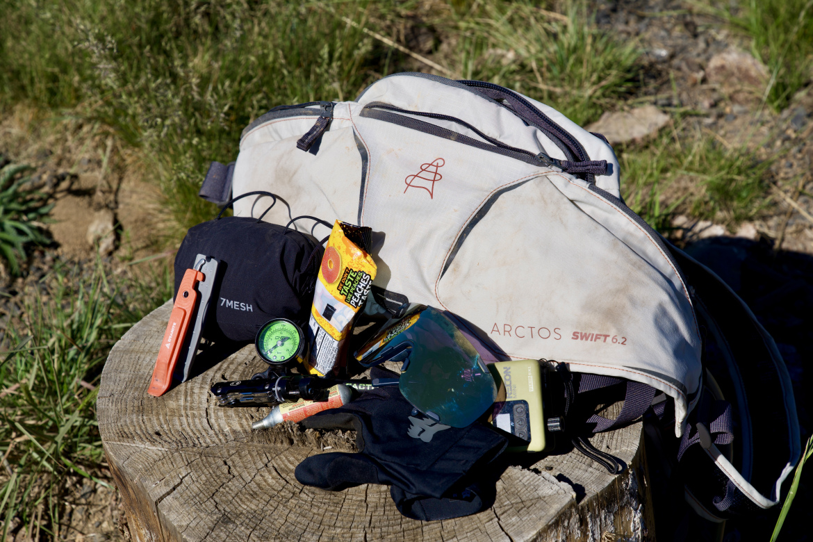 A variety of outdoor gear is displayed on a wooden stump, including a beige Arctos Swift backpack, a black 7Mesh item, a multi-tool, a compass, a snack, sunglasses, and a small electronic device. The background features green grass and a natural setting, suggesting a hiking or camping environment.