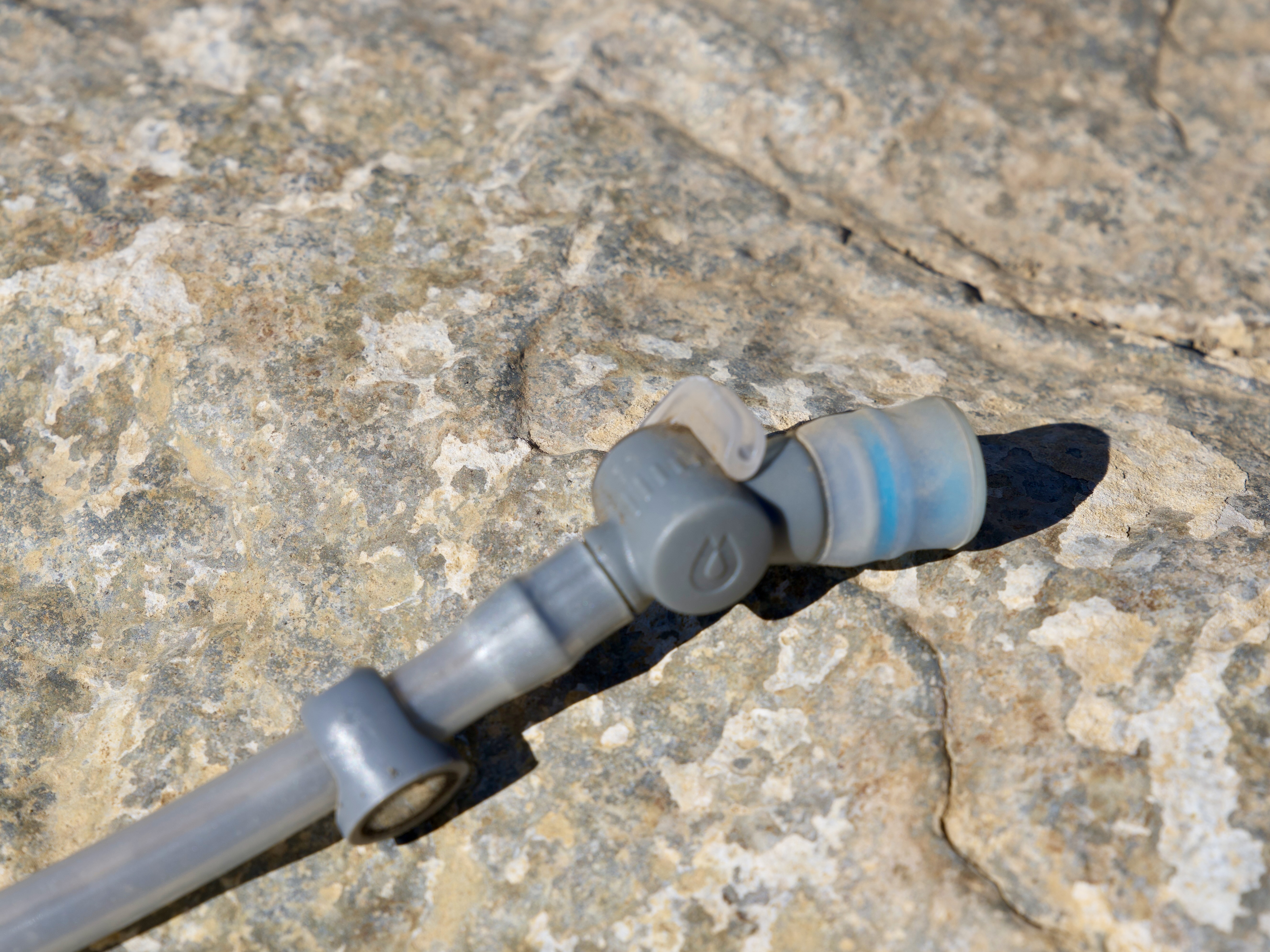 A close-up view of a gray flexible pipe or tubing resting on a textured rock surface, with a connection point visible and a small clear section at one end. The background features a natural rock texture with variations in color and pattern.