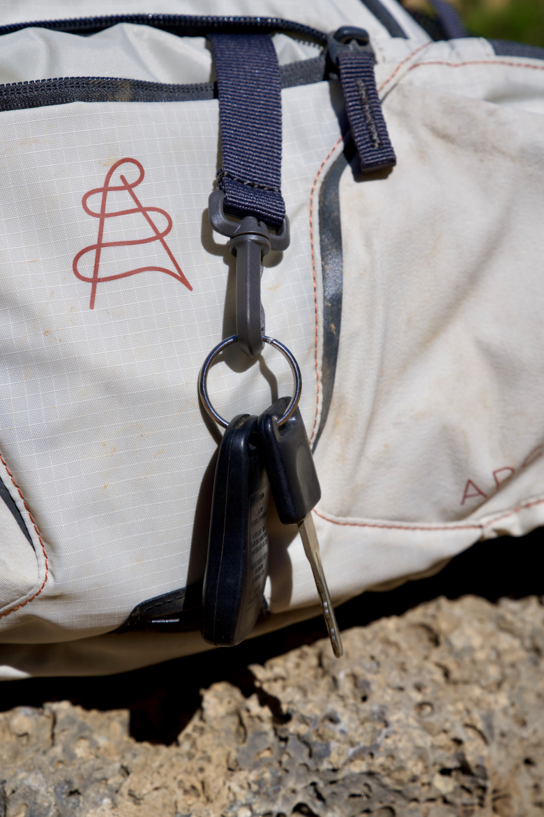 A close-up image of a light-colored outdoor backpack featuring a logo and a car key attached to a keyring clipped onto a strap. The backpack shows signs of wear, with some dirt visible on the fabric. The background includes a natural rocky surface.