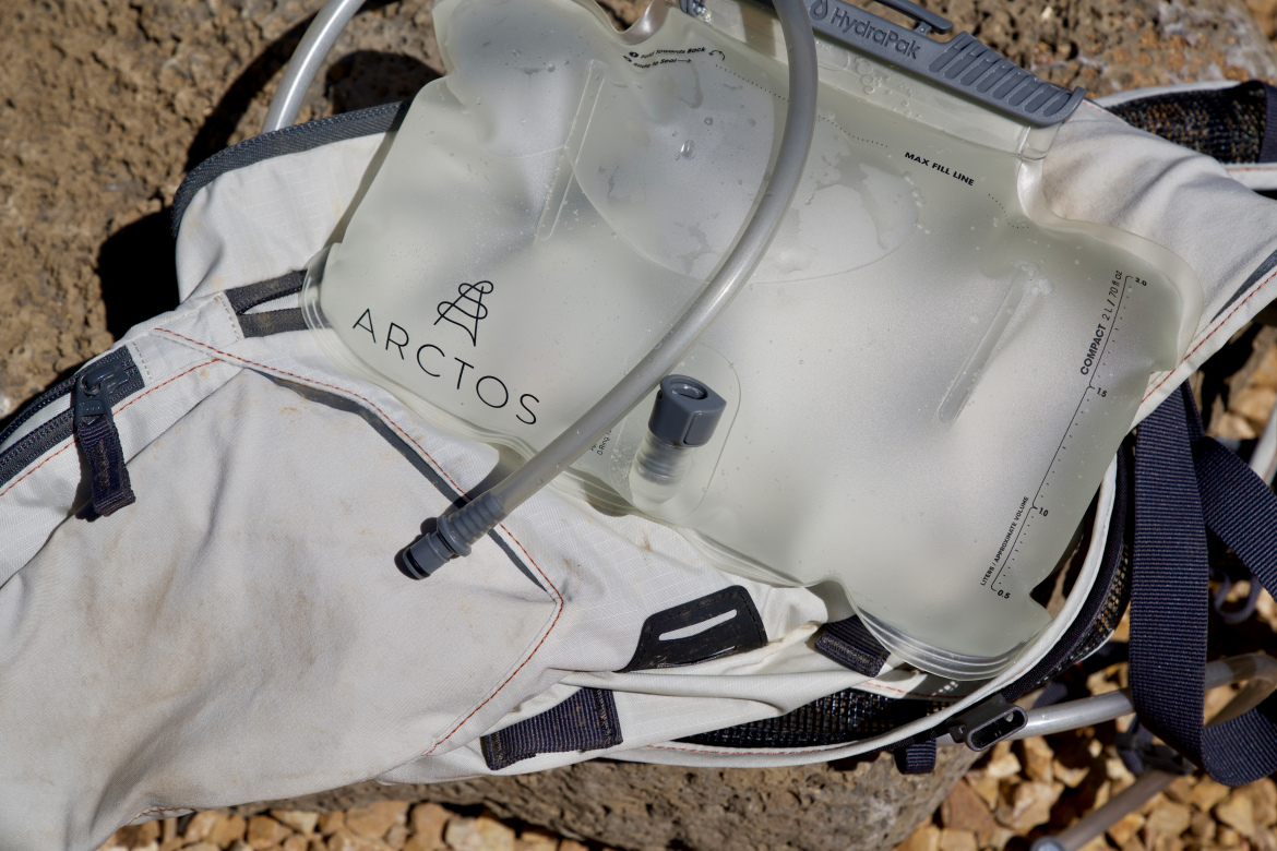 A close-up of a hydration bladder labeled "ARCTOS" resting on a white backpack, with a hose attached. The background shows rocky terrain under bright sunlight.
