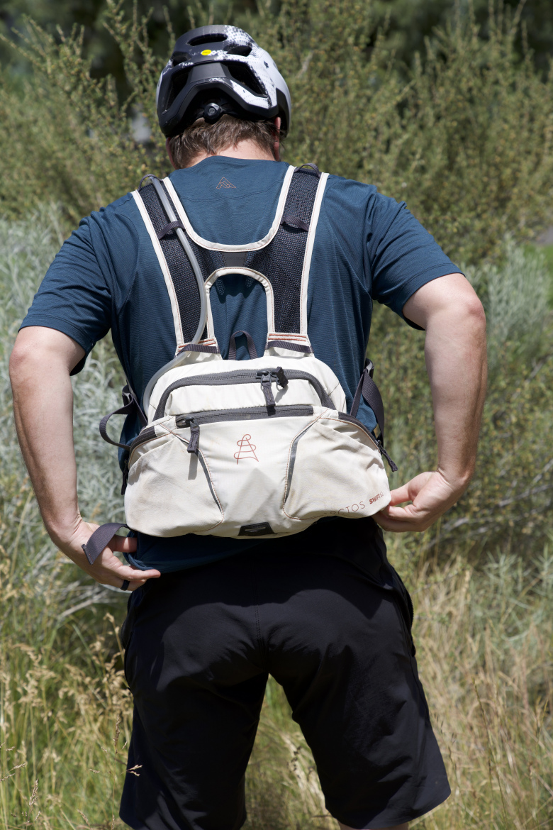 A person wearing a helmet and a blue athletic shirt stands with their back to the camera, adjusting a light-colored backpack with multiple compartments over their shoulders. The background features green foliage and tall grass, indicating an outdoor setting suitable for biking or hiking.