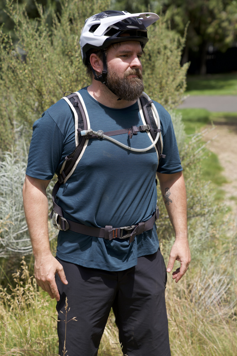 A man with a beard is standing outdoors, wearing a helmet and a hydration backpack. He is dressed in a short-sleeve blue shirt and dark shorts, surrounded by greenery and tall grass. The background includes a pathway and shrubs, suggesting a mountainous or trail setting.