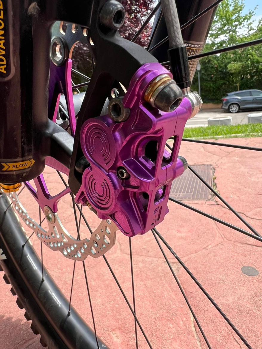Close-up view of a Hustok Race purple hydraulic disc brake caliper mounted on a mountain bike wheel, with a perforated metal rotor and surrounding bike components visible. The background includes greenery and a paved road, suggesting an outdoor setting.