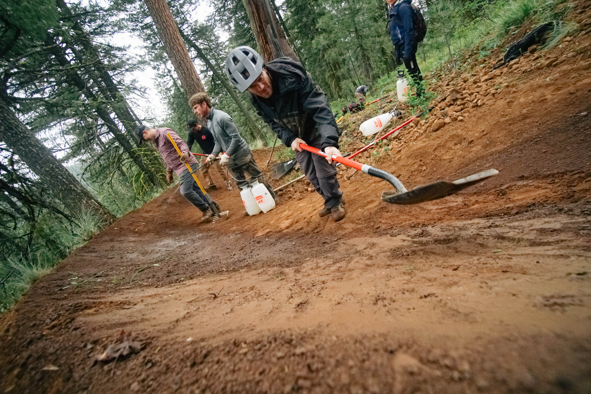 A group of people working together to build a dirt trail at ponderosa state park. They are using various tools, including shovels and rakes, while surrounded by tall trees and vegetation. Some participants are wearing helmets and gloves for safety, and there are water containers placed nearby.