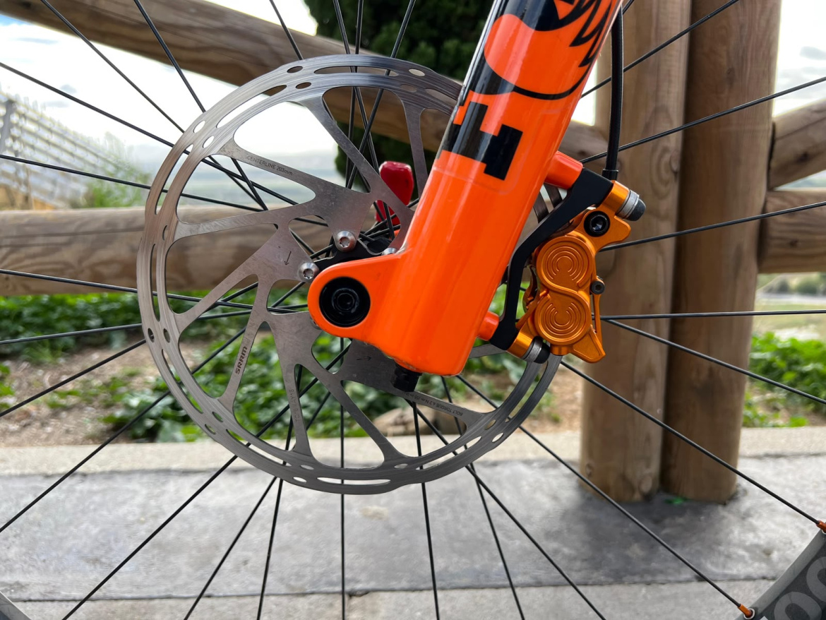 Close-up image of a bicycle's front wheel, showcasing a shiny metallic disc brake and orange Hustok brake caliper attached to the frame. The background features wooden fencing and greenery, suggesting an outdoor setting. The wheel spokes are visible, emphasizing the disc brake system.