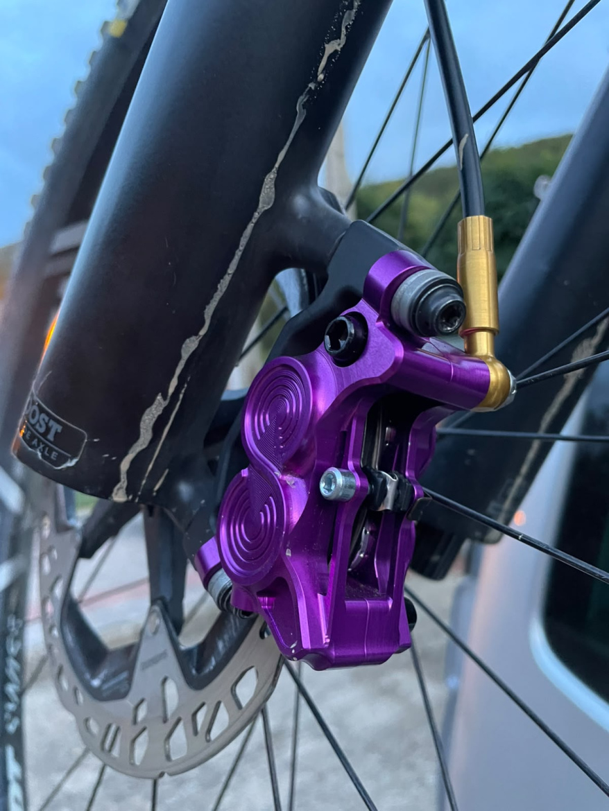 Close-up view of a Hustok Sprint purple bicycle brake caliper attached to a black fork, with a metallic brake line and disc brake visible in the background. The bike's frame shows some wear marks, and the background includes a blurred silver vehicle and natural scenery.