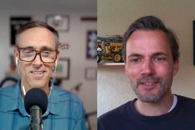 Two men appear in a split-screen video call. The man on the left has short hair and is wearing glasses and a blue plaid shirt, sitting in front of a microphone. The man on the right has medium-length hair and a beard, wearing a dark sweater, with a background featuring a shelf displaying a model vehicle and a framed picture. Both are smiling and engaged in conversation.