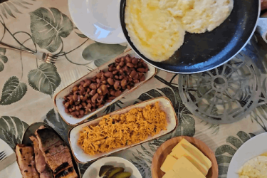 A person is serving scrambled eggs from a frying pan onto a plate, while a variety of food items are displayed on a table. The spread includes black beans, rice, pickles, tortillas, cheese, and slices of meat, all surrounded by a patterned tablecloth with leaf designs. Vashlovani National Park Adventure Loop mountain bike trail.