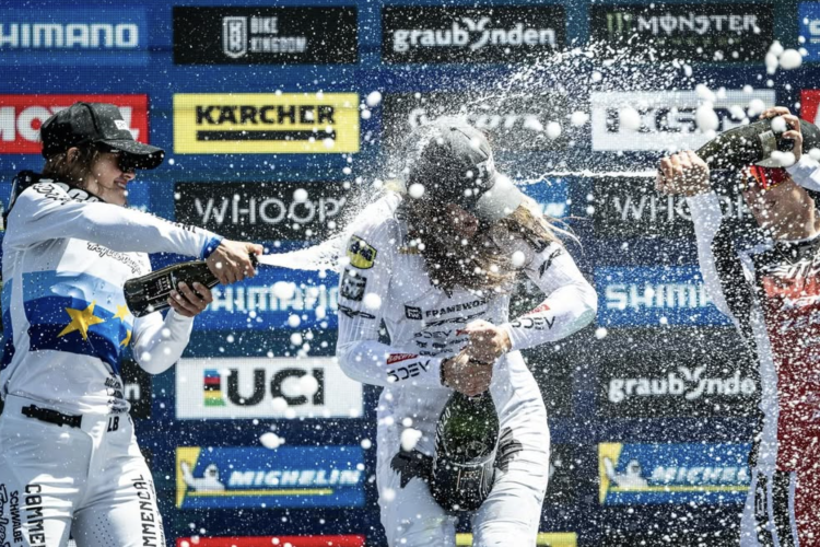 Three women in racing suits celebrating on a podium, spraying champagne and having fun. The background features sponsor logos and a blue backdrop, capturing a moment of joy and victory.