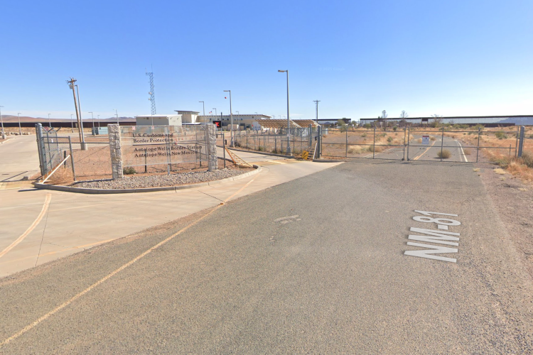 Exterior view of the Antelope Wells Border Station in New Mexico, featuring a gated entrance with a sign for U.S. Customs and Border Protection. The surrounding area is arid with sparse vegetation, and the sky is clear.