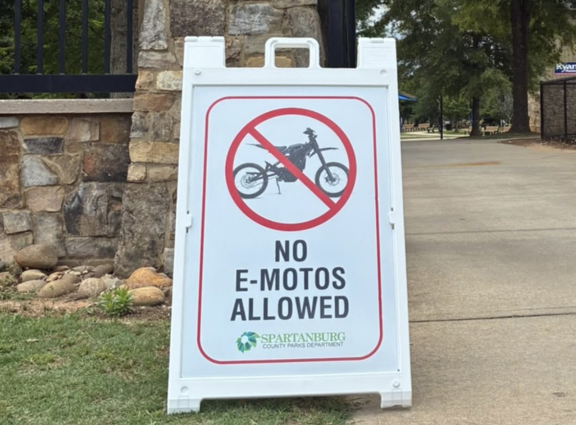 A white sandwich board sign stands on a pathway, displaying a graphic of a motorcycle crossed out in red, accompanied by the text "NO E-MOTOS ALLOWED" in bold black letters. The sign is identified as belonging to the Spartanburg County Parks Department. In the background, there are trees and grass, indicating a park setting.