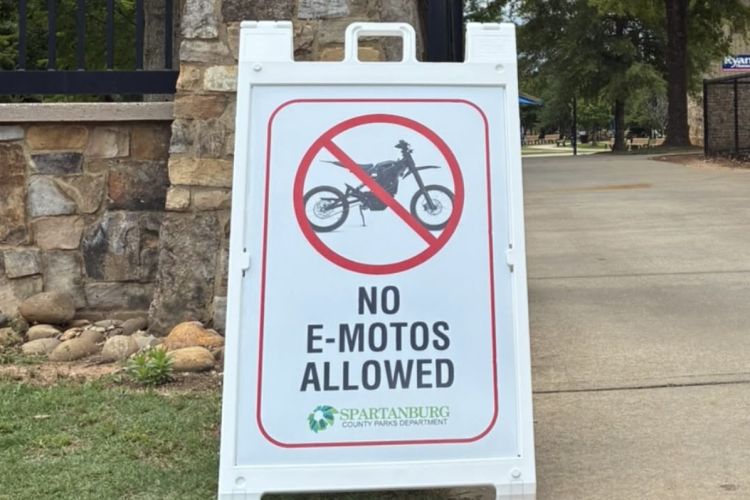 A white sandwich board sign stands on a pathway, displaying a graphic of a motorcycle crossed out in red, accompanied by the text "NO E-MOTOS ALLOWED" in bold black letters. The sign is identified as belonging to the Spartanburg County Parks Department. In the background, there are trees and grass, indicating a park setting.
