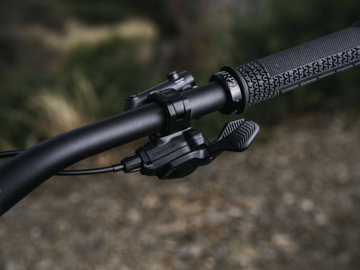 Close-up of the new Shimano Deore M6200 shifter. The shifter is positioned on the left side, with a wire connected to it. The background is blurred, emphasizing the handlebar setup with muted natural colors.