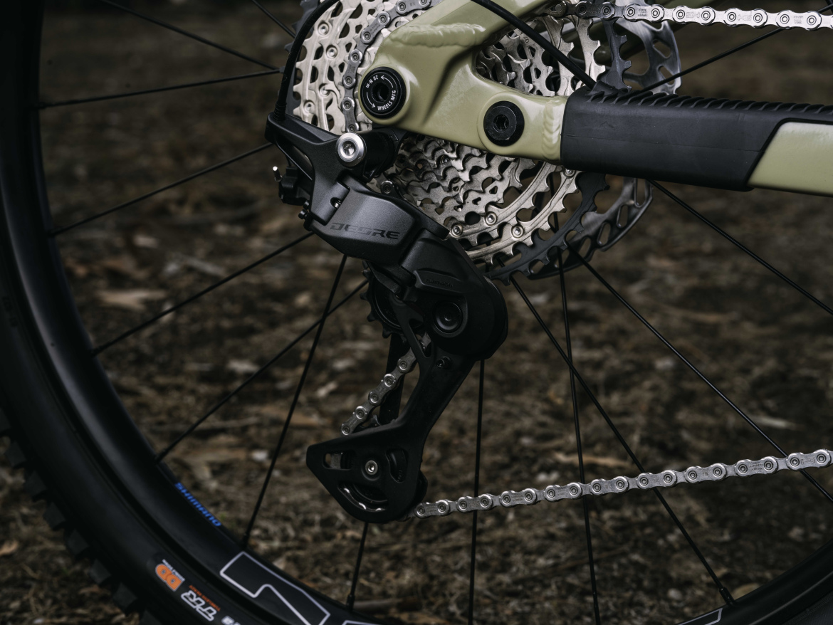 Close-up view of a bicycle's rear derailleur and cassette, Shimano's new mechanical Deore, showcasing the intricate gear mechanism and chain in a natural outdoor setting. The derailleur is positioned on the right side, with a black finish contrasting against the light-colored cassette. The background features blurred foliage and ground, emphasizing the focus on the bike components.