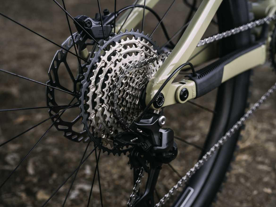 Close-up view of the new Shimano M7200 Deore Cassette, showcasing the intricate metal gears and chain, set against a blurred natural background. The bicycle is a modern design with a muted color frame.