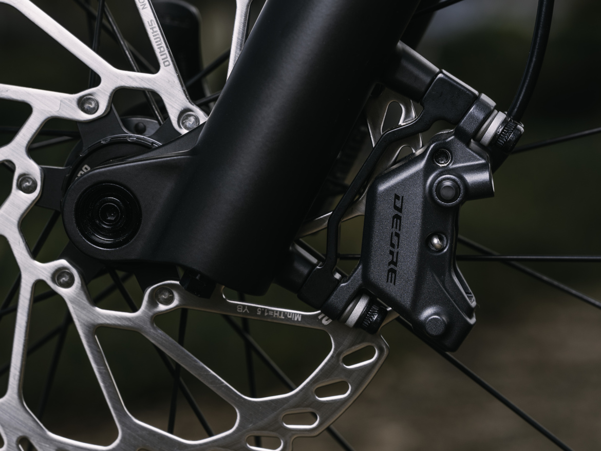 Close-up view of the new Shimano Deore M6220 brakes, featuring a metallic rotor with distinctive cutouts and a sleek caliper. The image highlights the intricate components, including the mounting mechanism and brake line, set against a blurred outdoor background.