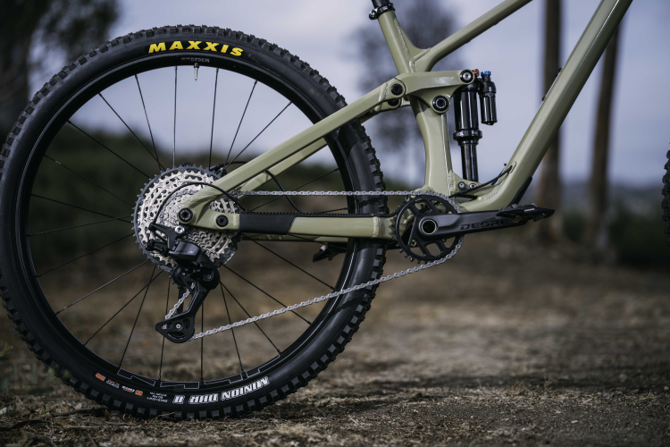 A close-up view of a mountain bike's drivetrain, featuring a matte olive green frame, a Shimano Deore crankset, and a detailed gear system. The image displays a Maxxis tire and a rear derailleur, with a blurred outdoor background that suggests a rugged terrain.
