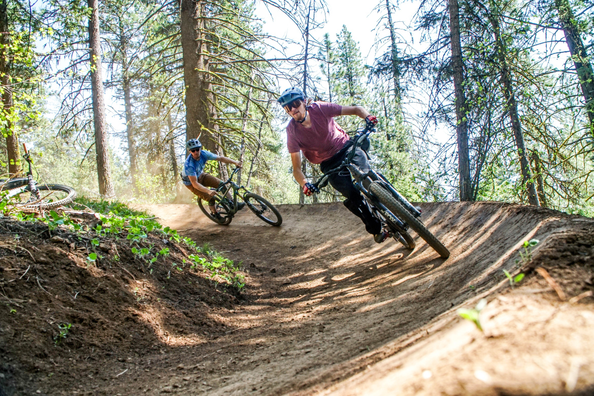Two mountain bikers navigate a dirt trail at ponderosa state park, leaning into sharp turns as dust rises behind them. One rider is wearing a red shirt and a helmet, while the other is dressed in a blue shirt. The scene captures the thrill of mountain biking amidst tall trees and greenery, with sunlight filtering through the branches.