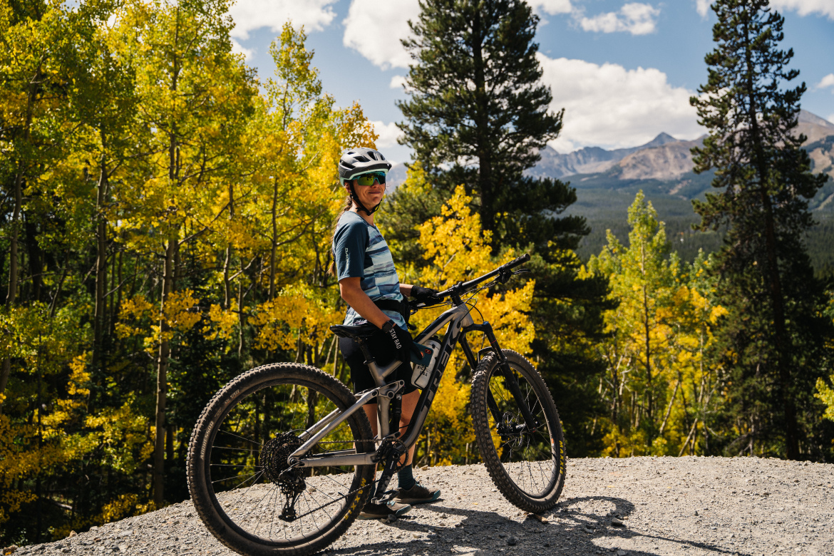 A person stands beside a mountain bike off the Aspen Alley trail, surrounded by vibrant yellow autumn foliage and tall trees. The backdrop features distant mountains under a partly cloudy sky. The cyclist is wearing a helmet and sunglasses, dressed in a casual outdoor outfit, and appears to be enjoying the scenic view.