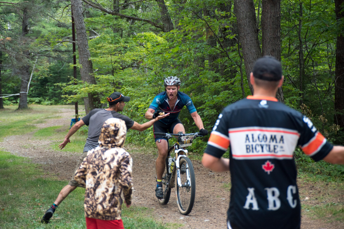 A mountain biker in a blue and black jersey rides on a dirt trail, making an intense expression while being cheered on by spectators. One person, wearing a cap, reaches out towards the biker, while another in a camouflaged hoodie and a red shirt observes the event. The scene is set in a wooded area with green trees in the background.