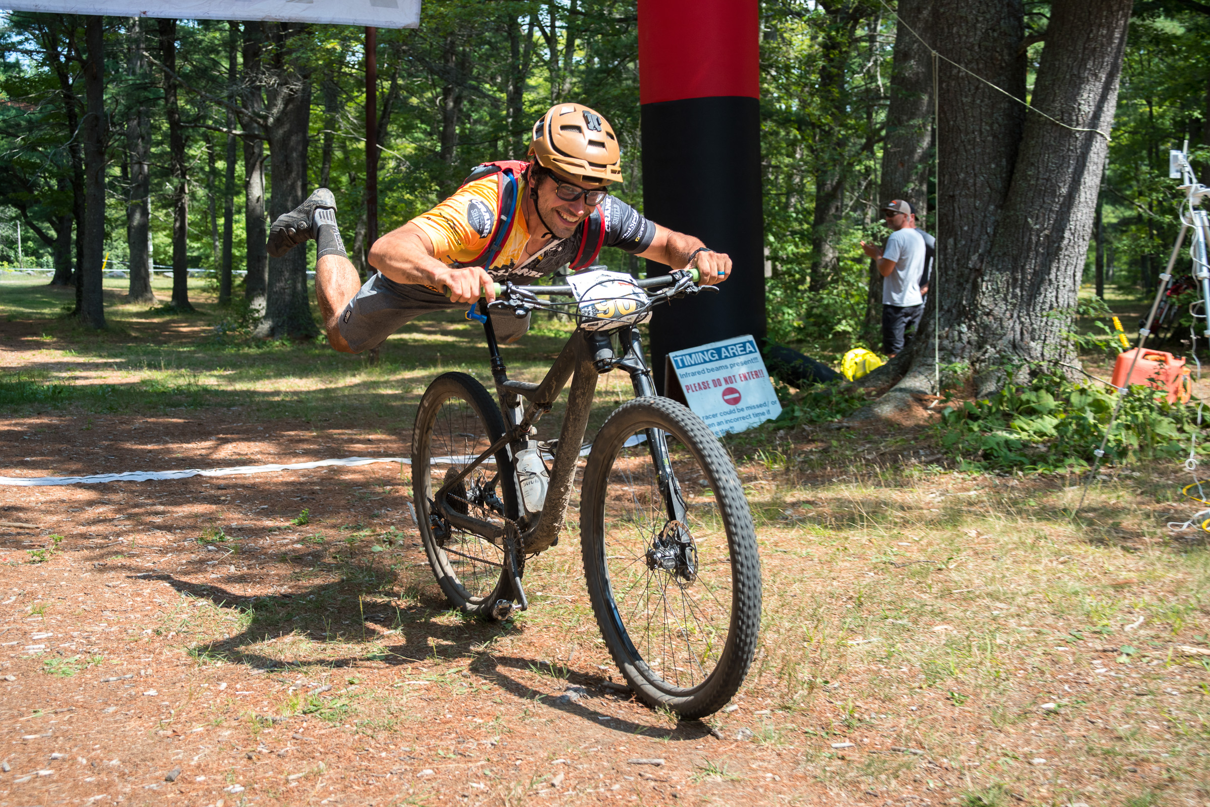 A mountain biker performing a stunt while racing through a wooded area, with one foot off the pedals and arms extended forward. In the background, spectators watch near a timing area with a sign, and trees surround the scene. The biker is wearing a helmet and colorful cycling gear.