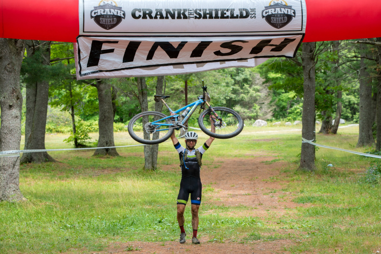 A cyclist triumphantly raises their mountain bike overhead at the finish line of a race, celebrating the achievement. The backdrop features a lush green landscape with trees, and a large "FINISH" banner hangs above. The cyclist is wearing a helmet and a race outfit, showing signs of mud from the trail.