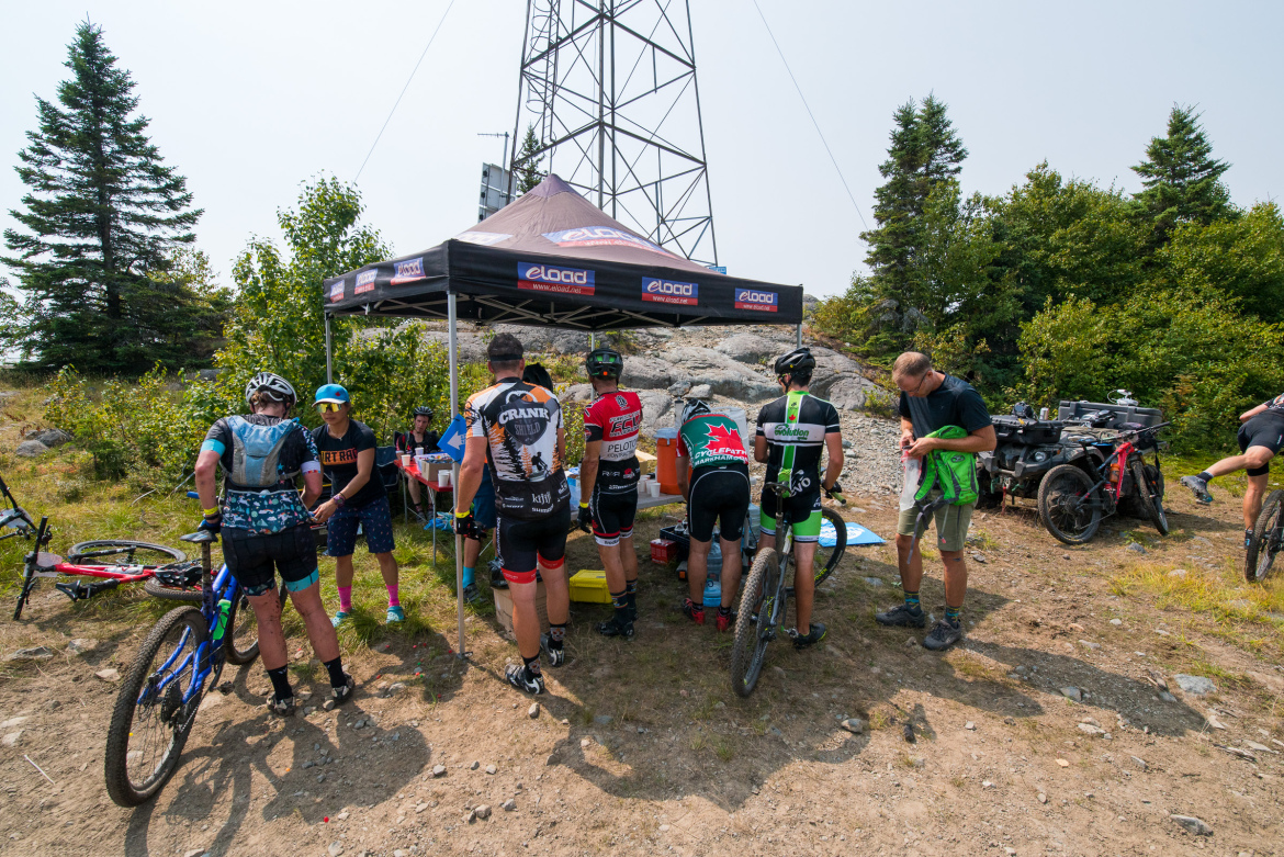 A group of mountain bikers gather around a tent set up near a wooded area. They appear to be taking a break, with some cyclists adjusting their gear and others engaging in conversation. Various mountain bikes are scattered nearby, and a supportive atmosphere is evident as they recharge in a natural landscape. A communications tower is visible in the background, adding a touch of rugged adventure to the scene.