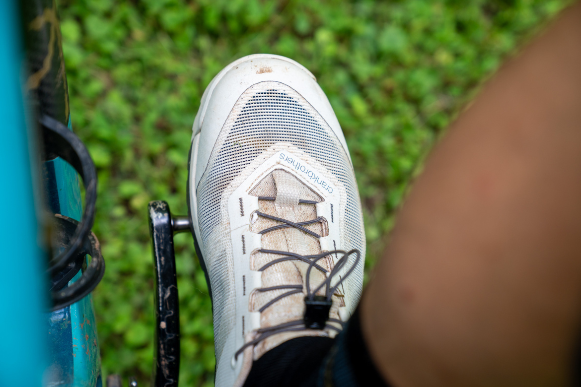 A close-up view of a cyclist's foot wearing a light-colored cycling shoe with visible dirt, resting on a black bicycle pedal. The background features green grass.