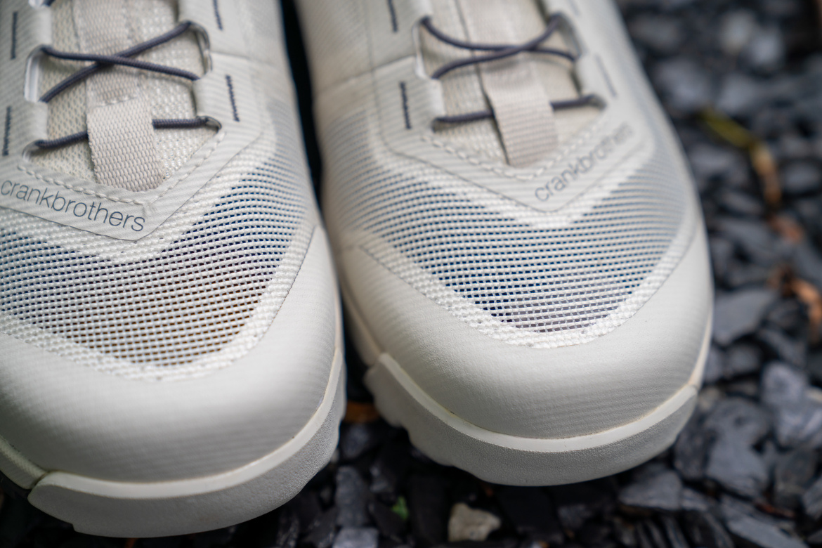 Close-up view of light-colored cycling shoes featuring mesh material and detailed lacing, resting on a gravel surface. The shoe has the brand name "Crankbrothers" visible on the side.