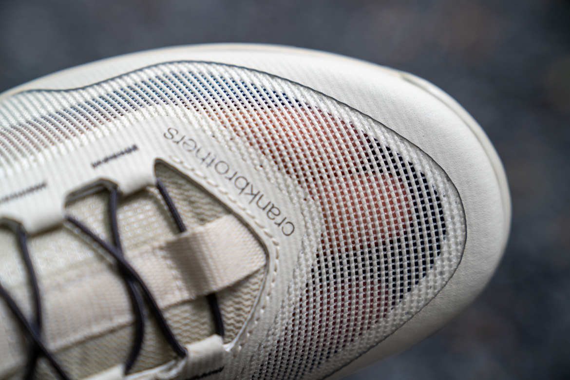 Close-up view of a lightweight athletic shoe with a textured upper featuring mesh panels and elastic lacing. The shoe is primarily white with subtle color variations and branding visible on the side.