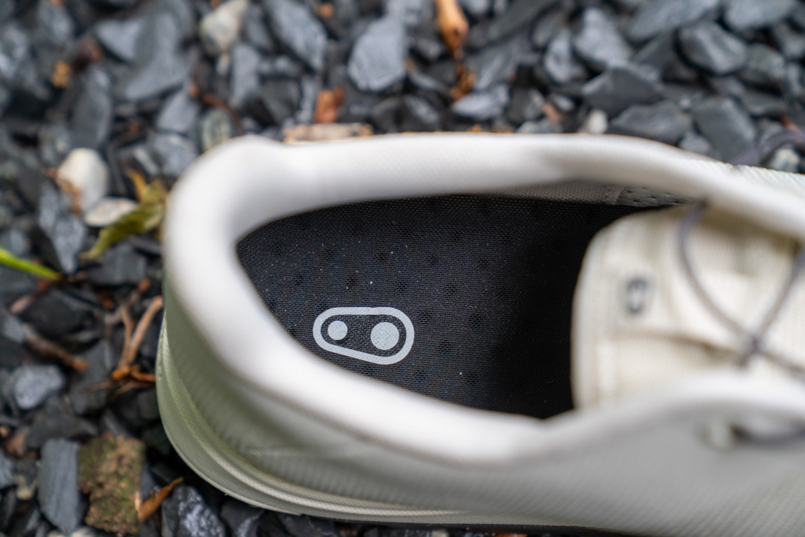 Top view of a light-colored sneaker resting on a bed of small, dark stones. The interior shows a black insole featuring a distinctive logo, with details of the shoe's structure visible around the edges.