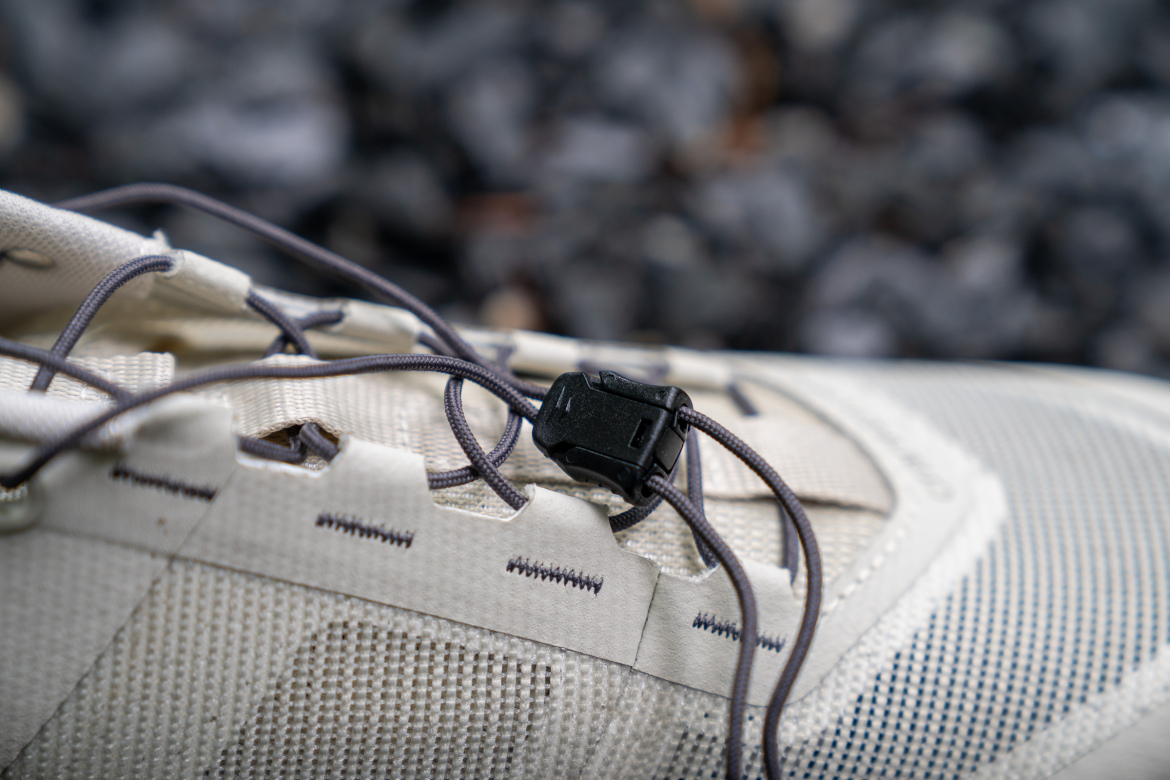 Close-up of a light-colored athletic shoe featuring a flexible lacing system with a locking mechanism. The shoe is set against a textured, rocky background.