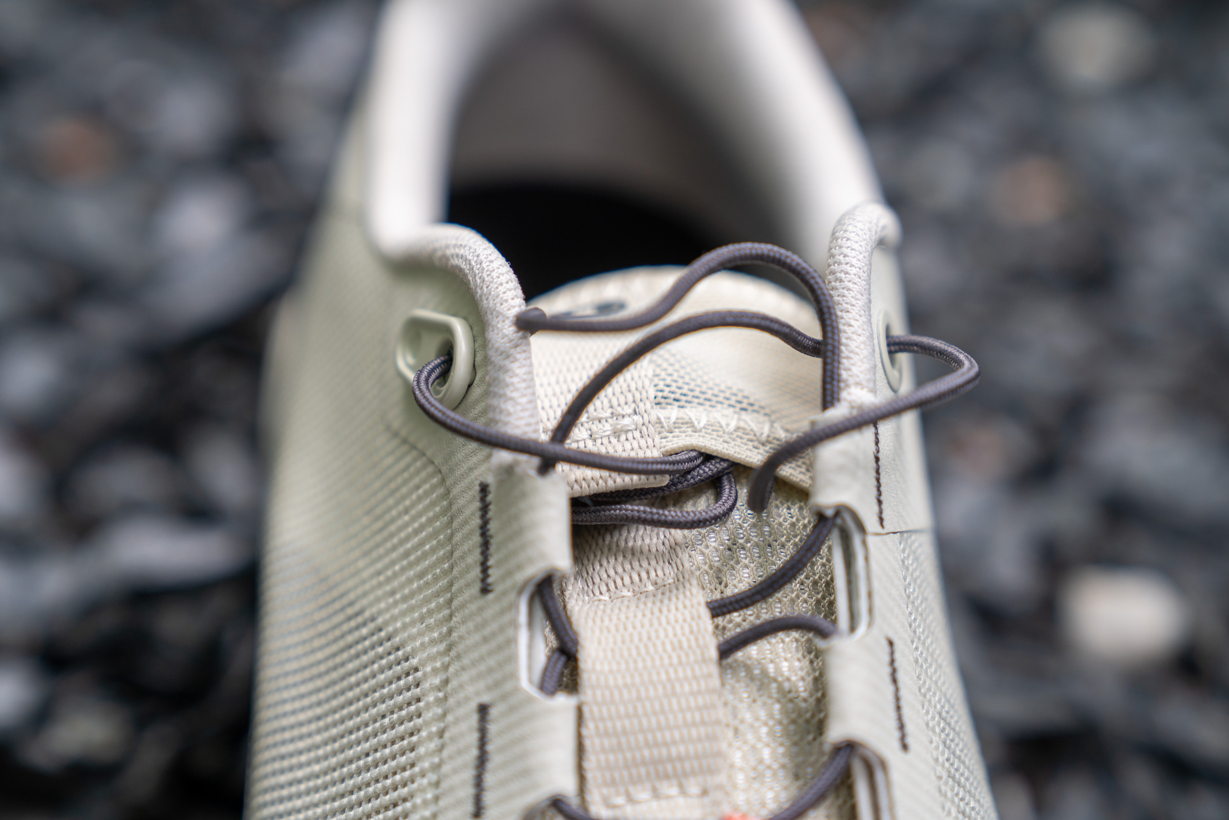 Close-up of a beige athletic shoe with laces, focusing on the top section where the laces are threaded through eyelets. The background features a textured surface, enhancing the shoe's details.