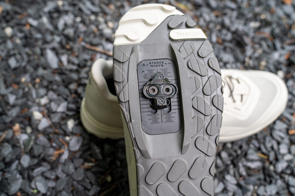 Close-up view of the sole of a light-colored athletic shoe, showcasing a clip mechanism for attaching to a pedal system. The tread pattern is visible, along with markings for stance and width adjustments. The background consists of small, dark gravel.